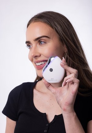 Stylpro-Gesichtsgerät, weiß mit metallischen Akzenten, geschwungene Form, lila Licht, Taste an der Vorderseite, wird von einer Person nah am Gesicht gehalten.