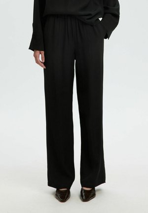Personne portant un pantalon noir ample, un haut noir avec une main dans la poche et des chaussures pointues foncées, debout devant un fond uni.