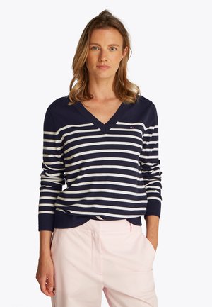 Femme portant un pull à col en V à rayures bleu marine et blanc avec un pantalon rose clair, debout avec une main dans la poche sur un fond uni.
