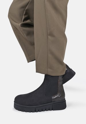 Black ankle boots with textured panel and thick ridged sole worn with brown cropped pants against a plain background.