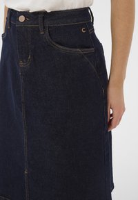 Tamsiai mėlynas džinsinis sijonas su matomu siuvimu, šoniniais kišenėmis ir aptempta juosmens linija. Turi sagų užsegimą ir tiesų siluetą.