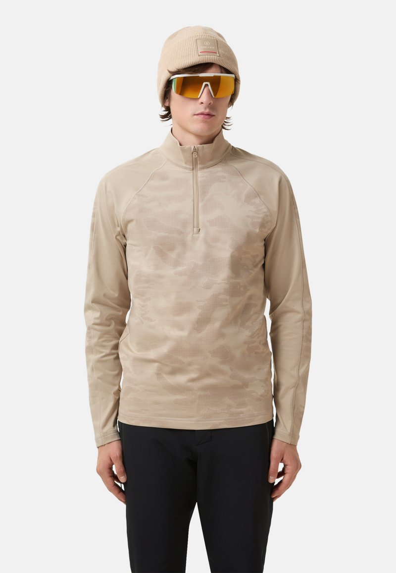 Beige Langarmshirt mit einem Viertel-Reißverschluss, mit einem Camouflage-Muster und glatter Textur. Getragen mit einer beigen Mütze und reflektierenden Sonnenbrillen.