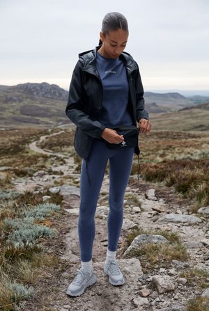 Kvinne justerer svart midjeveske på en steinete tursti, iført marineblå treningstøy og svart jakke, med fjellandskap i bakgrunnen.