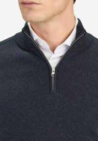 Sininen neulepusero, jossa on korkea kaulus ja vetoketjusuljenta. Kankaan pinta vaikuttaa sileältä, mutta siinä on hienoinen tekstuuri, ja se on kerroksittain valkoisen paidan päällä.