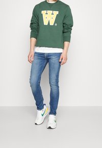 Muž oblečený v zelené mikině s žlutým logem "W", bílém tričku s krátkým rukávem, modrých skinny džínách a bílých teniskách s zelenými a žlutými akcenty.