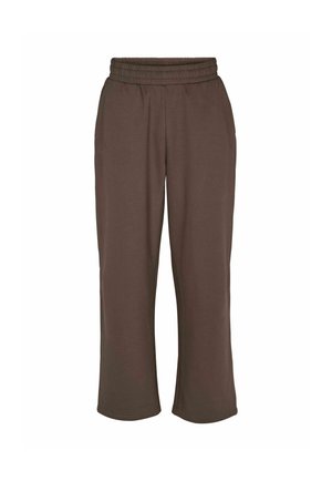Personne portant un pantalon de survêtement marron ample et un sweat assorti, les mains dans les poches, portant des baskets beige et blanches.