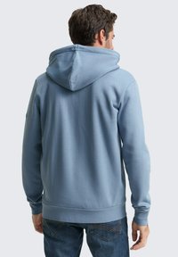 Lichtblauwe hoodie met een grote verstelbare capuchon, ribbelboorden en zoom, en een zachte textuur. G gedragen met blauwe spijkerbroek.