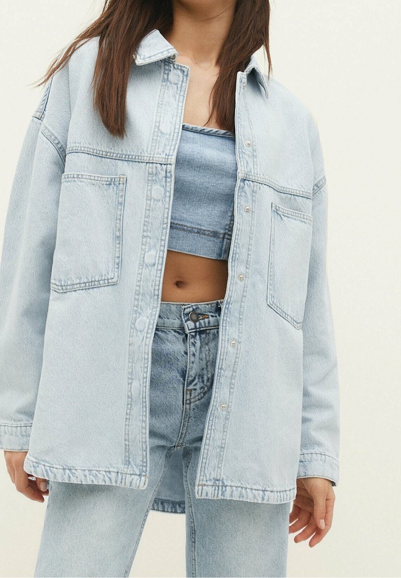 Light blue denim jacket with a relaxed fit, featuring a pointed collar, two front pockets, and visible stitching details.