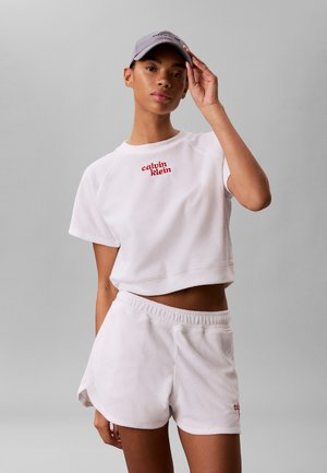 Kobieta w białym crop topie i szortach z frotté Calvin Klein z czerwonym logo, poprawiająca szarą czapkę na jednolitym, szarym tle.