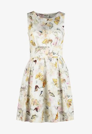 Kobieta spacerująca w białej sukience bez rękawów z motywem motyli, czarnych przezroczystych rajstopach, beżowych botkach do kostki i z czarnymi paskami plecaka.