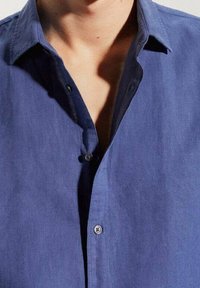 Camicia blu con colletto strutturato, caratterizzata da una texture liscia e un unico bottone visibile nella patta.