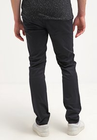 Schwarze Slim-Fit-Hosen mit glatter Textur und dezentem Glanz, kombiniert mit weißen Sneakers. Ansicht von hinten, die das taillierte Design zeigt.