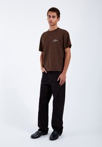 Junger Mann steht mit den Händen in den Taschen, trägt ein braunes T-Shirt mit weißem Text und schwarze Hose vor weißem Hintergrund.