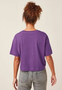 Fioletowy skrócony t-shirt wykonany z miękkiej bawełny, z krótkimi rękawami i luźnym krojem. Noszony z szarymi dżinsami z wysokim stanem.