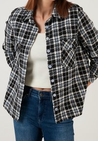Camicia a quadri bianca e nera con accenti blu, realizzata in tessuto texturizzato, con chiusura a bottoni, colletto e taschino sul petto.