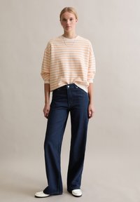 Przesadnie duży sweter w brzoskwiniowo-białe paski, zestawiony z ciemnoniebieskimi jeansami o szerokich nogawkach. Modelka nosi białe buty i srebrny naszyjnik łańcuchowy.