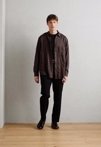 Camisa de botones marrón sobre una camiseta negra, combinada con pantalones negros y zapatos marrones. Tejido suave con un ajuste relajado.