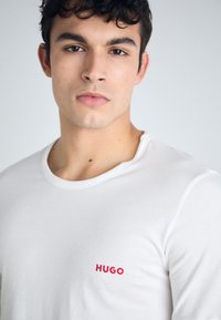 T-shirt en coton blanc avec un col ras du cou et un petit logo rouge "HUGO" sur la partie gauche de la poitrine. Texture simple et lisse.