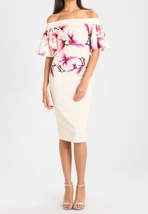 Woman wearing an off-shoulder white dress with pink floral print and ruffled sleeves, paired with silver high-heeled sandals.