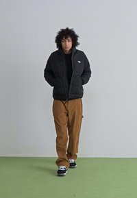 Chaqueta de plumas negra con cierre delantero, cuello alzado y parche de logo; combinada con pantalones de color beige y zapatillas negras sobre un suelo verde.