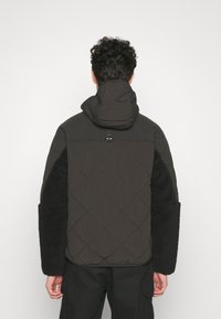 Veste matelassée noire avec capuche, présentant des manches en polaire texturée noire et un détail d'accroche à l'arrière. Design simple et fonctionnel.