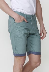 Shorts en denim verts avec des revers bleus, cinq poches et une coupe slim. Le tissu a une texture lisse avec un léger brillant.