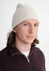 Crème ribgebreide beanie met een snug fit, gemaakt van zachte stof. Model draagt een chocoladekleurige zip-up sweater met een gestructureerd patroon.