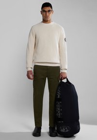 Beige gerippten Pullover, olivgrüne Cargo-Hosen, schwarze Tasche mit Logo-Print, getragen mit schwarzen Stiefeln, stehend vor einem neutralen Hintergrund.