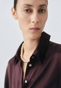 Camisa de satén en color burdeos con cuello, detalles de botones y un collar de perlas. Textura suave, diseño elegante y fondo neutro.