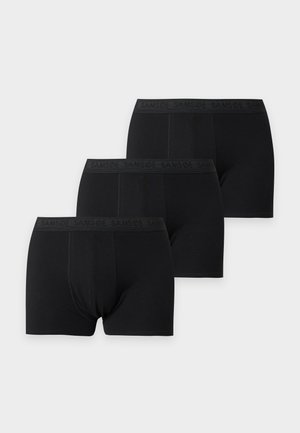 Homme debout pieds nus, bras croisés, portant un boxer noir sur un fond clair uni.