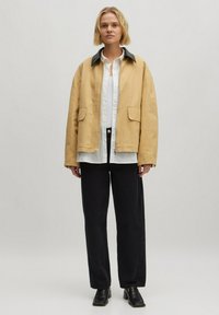 Beige Canvasjacke mit einem schwarzen Kragen, zwei Vordertaschen, über einem weißen Hemd und schwarzen Hosen getragen, kombiniert mit schwarzen Knöchelstiefeln.
