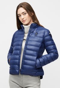 Γυναίκα με μακριά καστανά μαλλιά που φοράει μπλε μπουφάν με καπιτονέ ύφασμα πάνω από ένα ανοιχτό γκρι πουλόβερ με ζιβάγκο, με τα χέρια στις τσέπες.