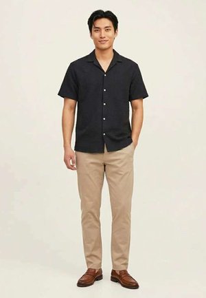 Hombre de pie, vistiendo una camisa negra de manga corta abotonada, pantalones beige y mocasines de cuero marrón, manos relajadas, fondo neutro.