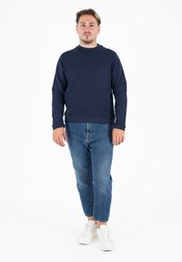 Felpa blu navy con collo a giro e cuciture sporgenti sulle spalle, abbinata a jeans azzurri a gamba affusolata e sneakers bianche, in piedi su uno sfondo bianco.