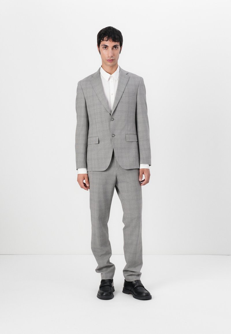 Completo da uomo grigio a scacchi con due bottoni, pantaloni coordinati, camicia bianca e scarpe in pelle nera. Tessuto morbido e vestibilità su misura.
