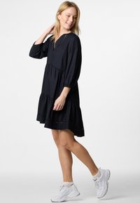 Schwarzes knielanges Kleid mit langen Ärmeln, einem Schlüssellochausschnitt und gestuften Rüschen. Getragen mit weißen Sportschuhen. Glattes, leichtes Material.