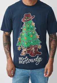 Tamsiai mėlynas marškinėlis su grafiniu dizainu, vaizduojančiu papuoštą Kalėdų eglutę, kaubojų skrybėlę, žibintus ir kaubojiškus batus. Užraše parašyta „Ho Ho Howdy“.