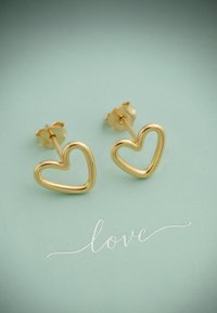 Goldene, herzförmige Ohrstecker mit einer glatten, polierten Oberfläche. Vor einem mintgrünen Hintergrund mit dem Wort "Liebe" in Schreibschrift präsentiert.
