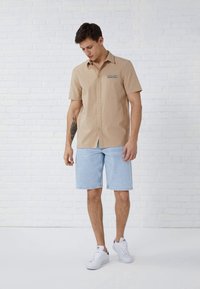 Beige short-sleeve button-up shirt with embroidered detail, paired with light blue denim shorts and white sneakers, standing against a white wall.