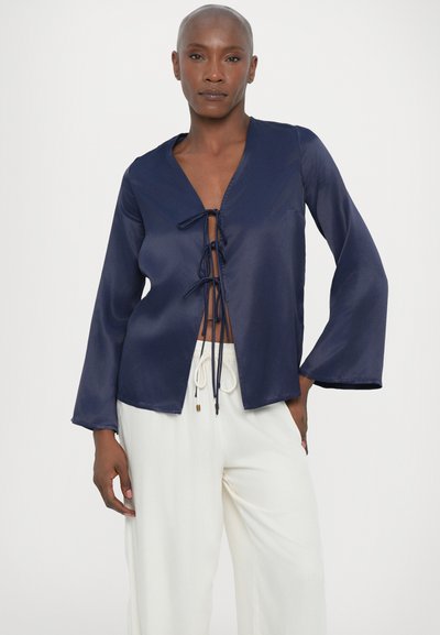 Blusa de satén azul marino con escote en V profundo, cierre frontal con cordones y mangas largas acampanadas. Presenta un diseño minimalista y una textura suave.