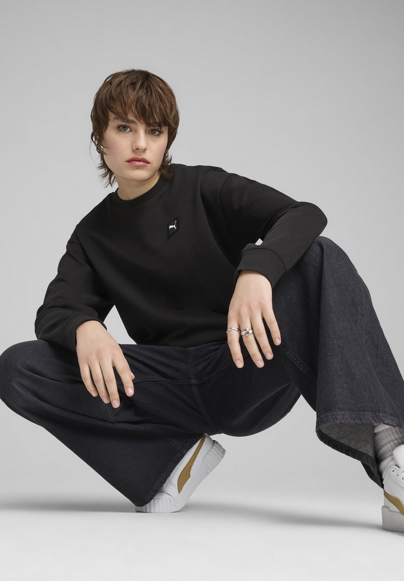 Schwarzer Sweatshirt mit Rundhalsausschnitt und Puma-Logo, kombiniert mit weit geschnittenen, dunklen Jeans, weißen Sneakers mit braunen Akzenten und silbernen Ringen.