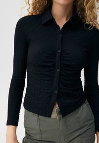 Chemise noire texturée à manches longues avec un devant à boutons et des détails froncés à la taille, associée à un pantalon vert olive.