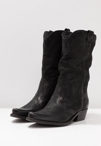 Para czarnych skórzanych butów kowbojskich z dekoracyjnym haftem i średniej wysokości obcasami na białej powierzchni.