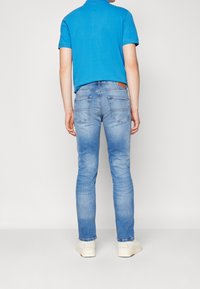 Jeans in denim azzurro chiaro con vestibilità slim, caratterizzati da tasche posteriori e un'etichetta in pelle marrone. Abbinati a una polo blu e sneakers bianche.