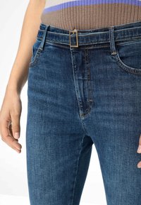 Jeans de mezclilla azul oscuro con corte ajustado, que presentan una hebilla metálica dorada y bolsillos cosidos, mostrando una textura suave y un ligero desgaste.