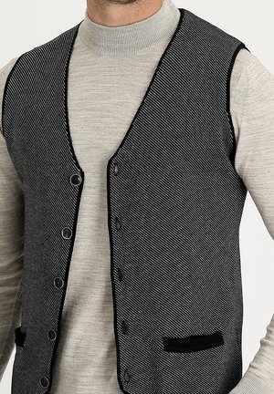 Homme portant un gilet texturé noir et blanc boutonné sur un pull à col roulé gris clair à manches longues.