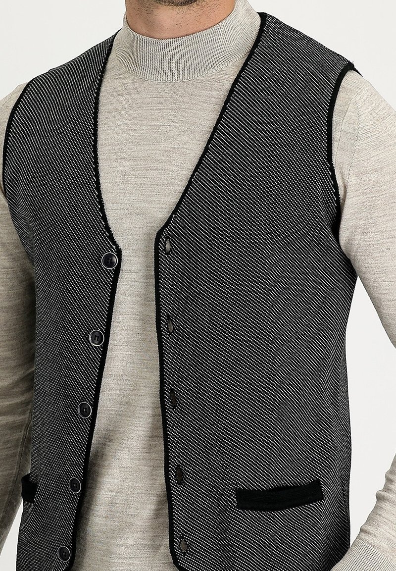Homme portant un gilet texturé noir et blanc boutonné sur un pull à col roulé gris clair à manches longues.