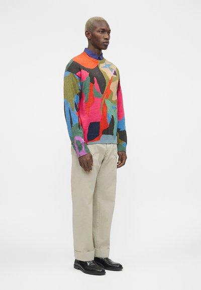 Άντρας με κοντά ξανθά μαλλιά που φοράει πολύχρωμο πουλόβερ με αφηρημένο μοτίβο, μπεζ παντελόνι και μαύρα παπούτσια, στέκεται πλαγίως.