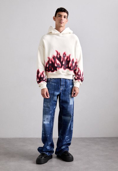 Hanorac cu glug�ă de culoare crem, cu un grafic de flacără roșie la tiv. Pantaloni din denim largi, cu imprimeu de ziar albastru. Pantofi cu toc înalt negri.