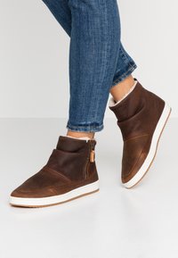 Brown leather ankle boots with a fold-over design, white rubber soles, and side zippers. Features a soft lining and minimal stitching details.
